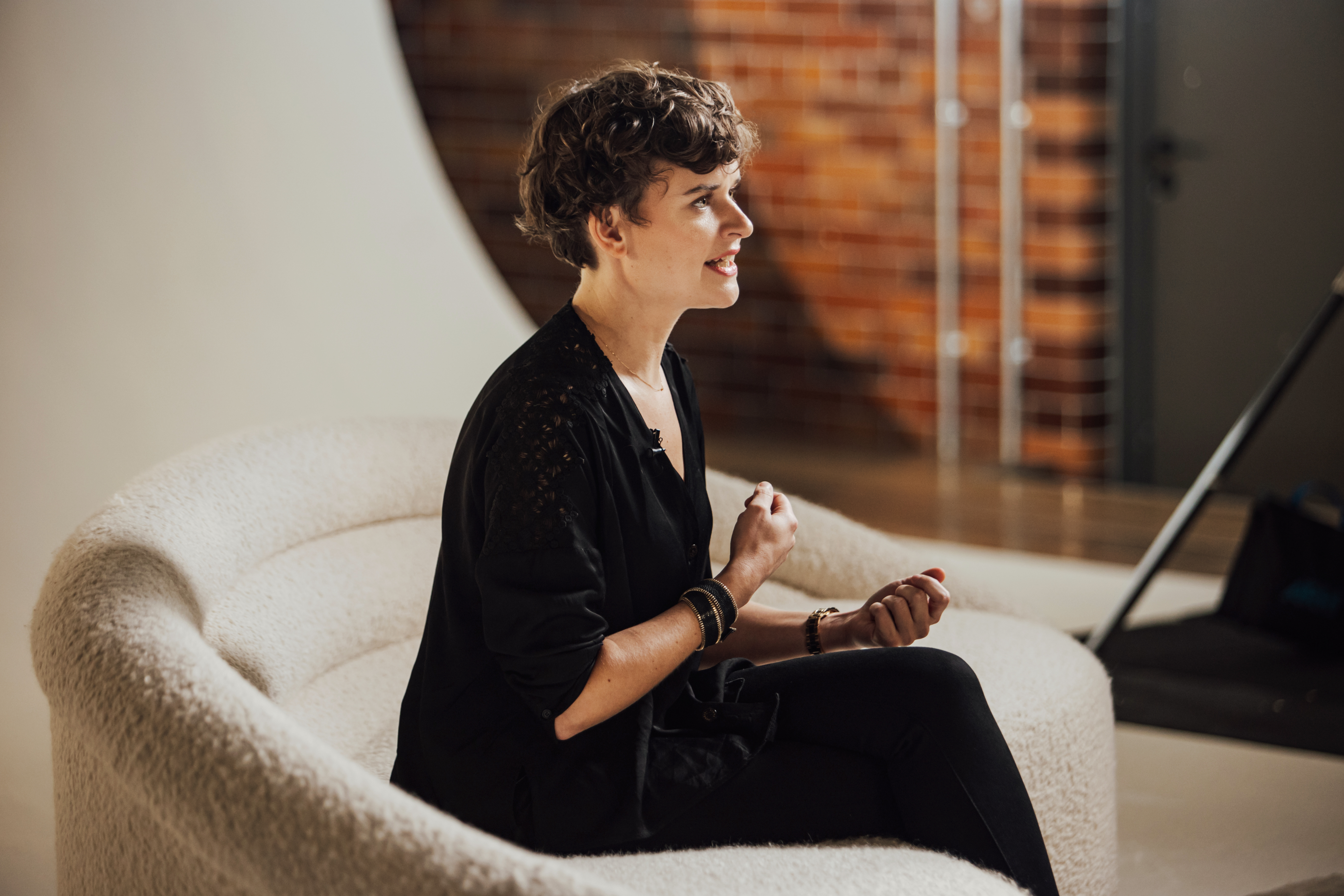 A person with short curly hair wearing a black outfit sits on a textured white couch in a studio with a brick wall background.