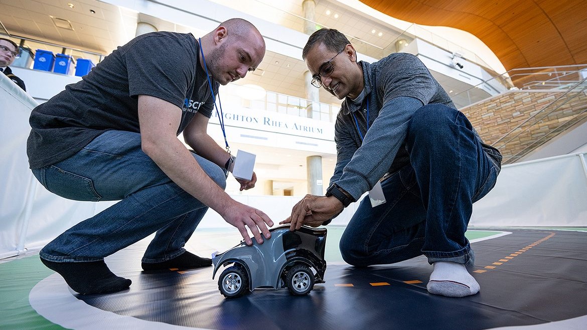 Two participants prepare a small autonomous vehicle on a track during an AWS DeepRacer robotics competition in a modern atrium setting.