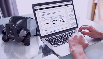 A person uses a laptop to train or program a small robotic car, with the laptop displaying a graphical interface for robotics or AI training applications.