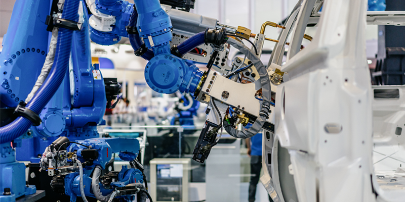 A blue industrial robotic arm equipped with computer vision technology is assembling or inspecting a white car frame in an automated automotive manufacturing facility.