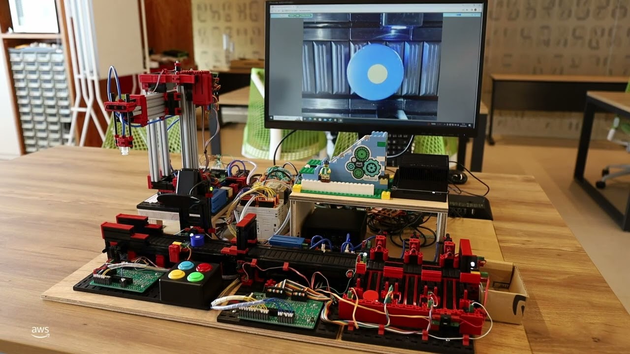 A robotics lab automation system setup on a table, featuring mechanical components, hardware circuits, and control buttons, with a computer monitor displaying a close-up view from the automated system in the background.