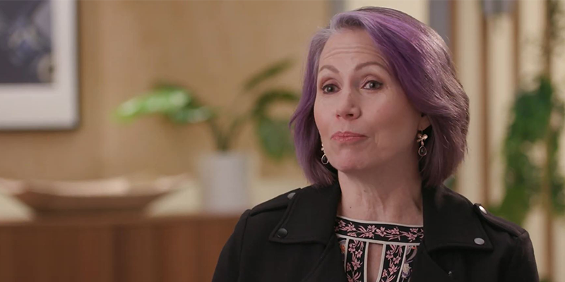 A woman with purple hair is speaking in an indoor setting with plants and art in the background.