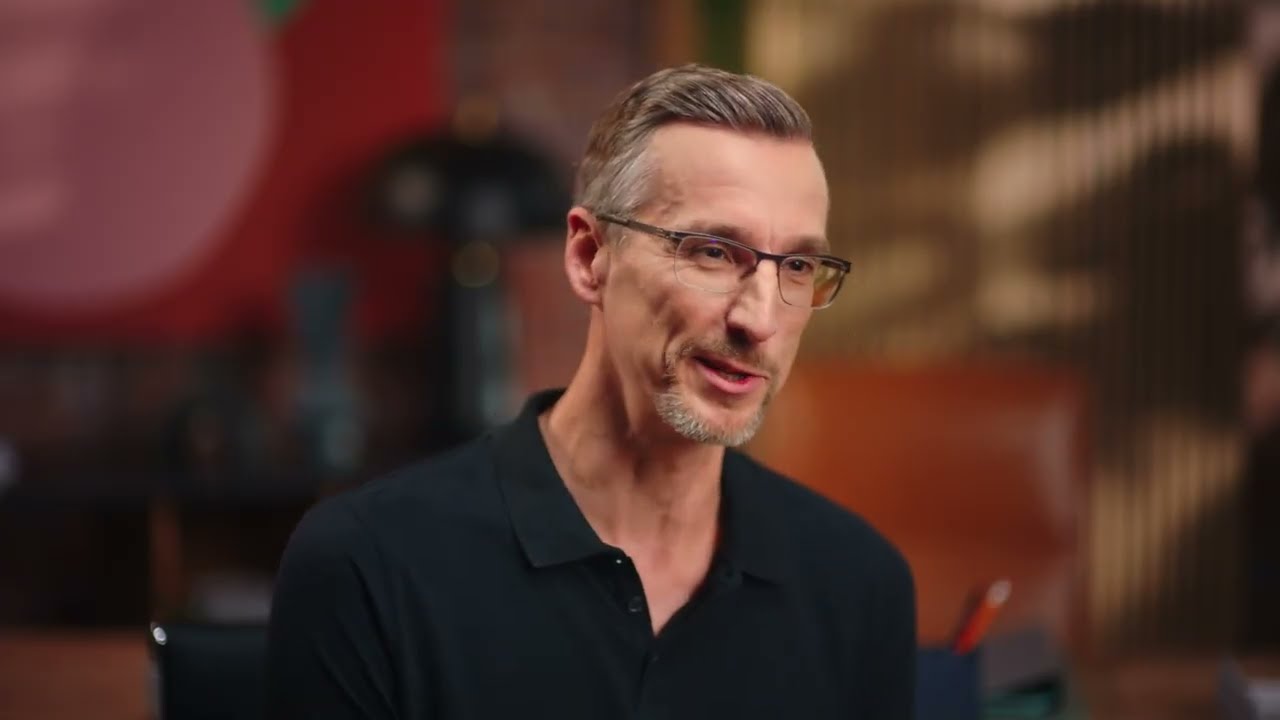 A professional wearing glasses and a dark polo shirt speaks in an office setting, suitable for topics related to security, identity, and compliance.