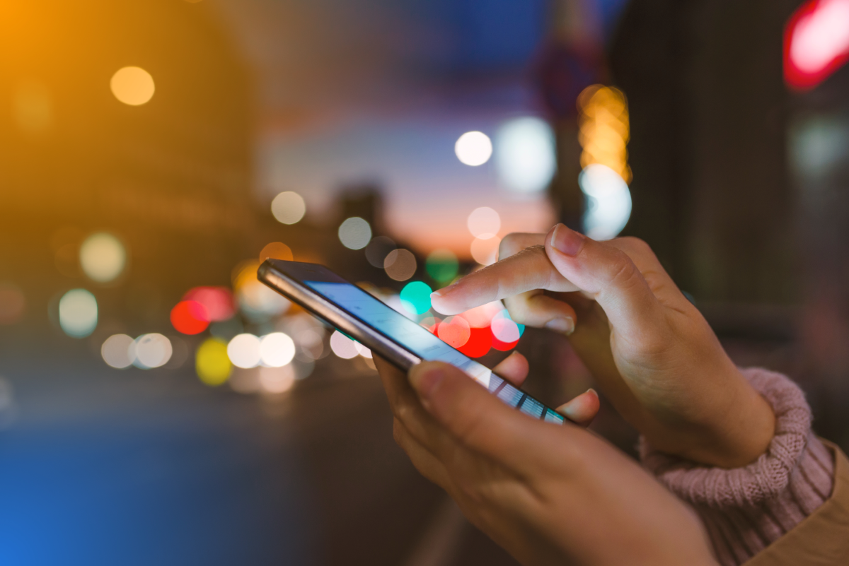 Close-up of hands using a smartphone on a city street at night with colorful blurred lights in the background.