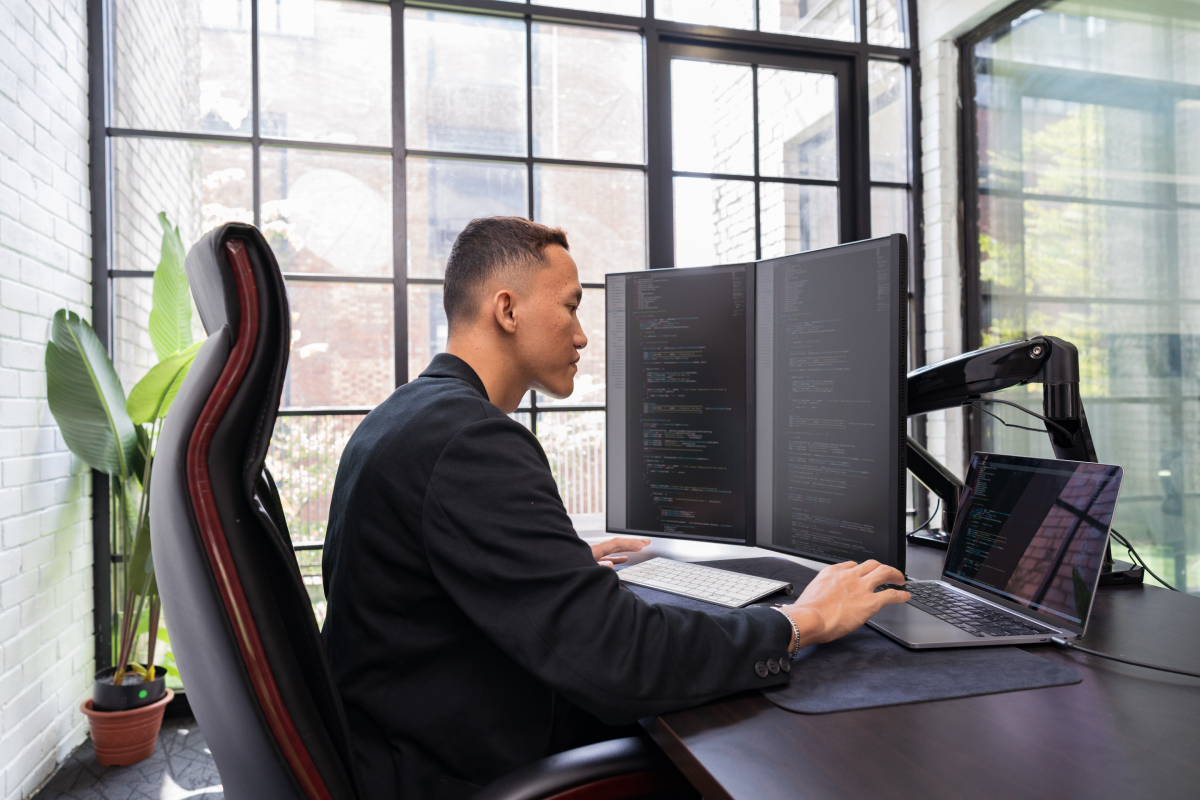 A software developer is working at a desk with dual monitors and a laptop in a modern office setting with glass windows. Computer code is visible on the screens.