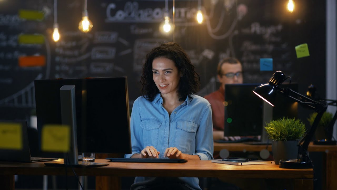 A person in a blue shirt working at a desk with a computer, surrounded by warm lighting and a chalkboard wall with collaborative notes in the background.