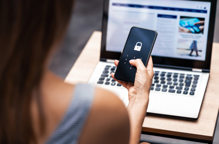 A woman holding a smartphone and entering a passcode to unlock it, with a laptop open in the background. The image represents digital security and identity protection.