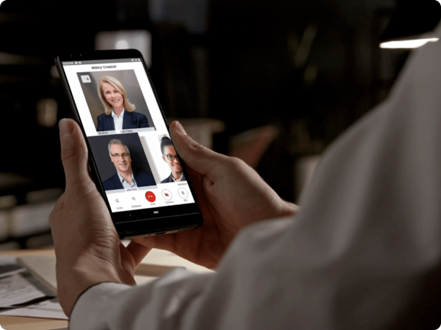 A person is holding a smartphone displaying a video conference call with three participants on the screen.