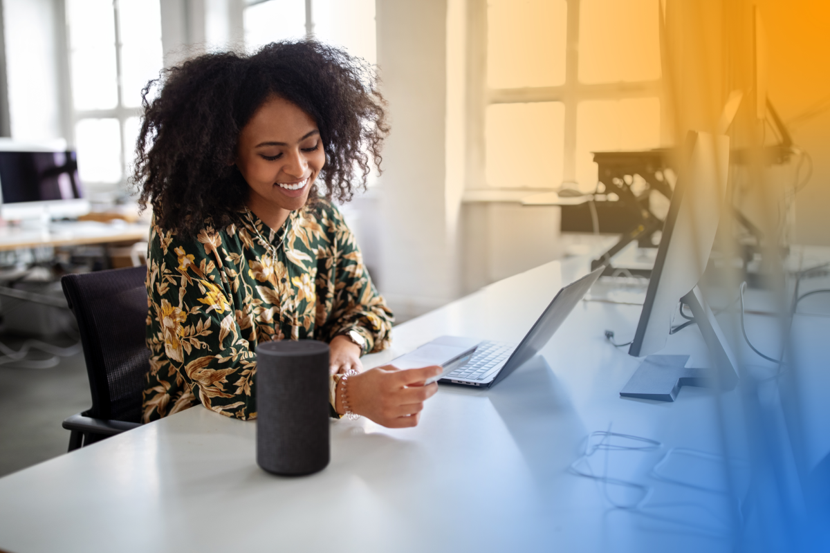 A woman is sitting at an office desk, entering credit card information on a laptop. She is smiling and appears to be working in a modern, bright workspace.