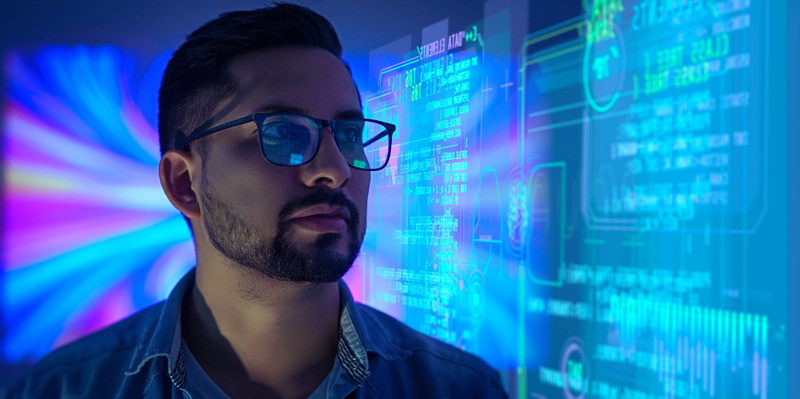 A man wearing glasses looks at a large digital screen displaying cybersecurity data, code, and visual analytics, representing security, identity, and compliance in a technology environment.