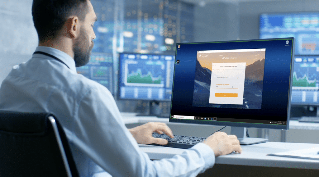 A man sitting at a desk in a modern office environment, typing on a keyboard while looking at a computer monitor displaying a login screen. Multiple screens with data and charts are visible in the background, emphasizing a secure or professional setting.