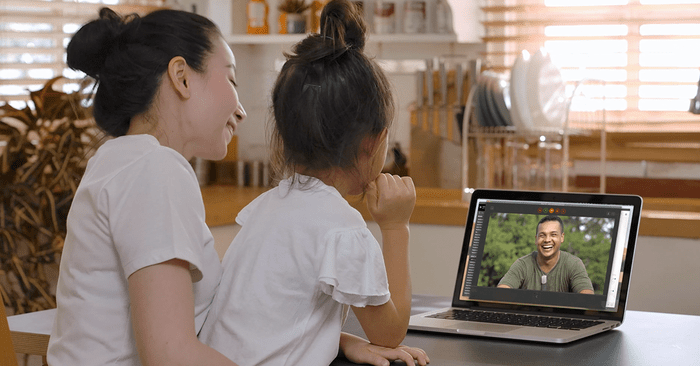 A woman and a girl are sitting together in a kitchen, smiling, while making a video call on a laptop. The screen shows a man smiling back at them.