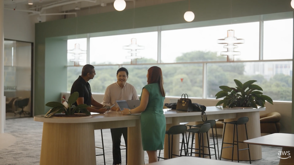 Three people standing around a modern high table in a bright office space with large windows and greenery outside.
