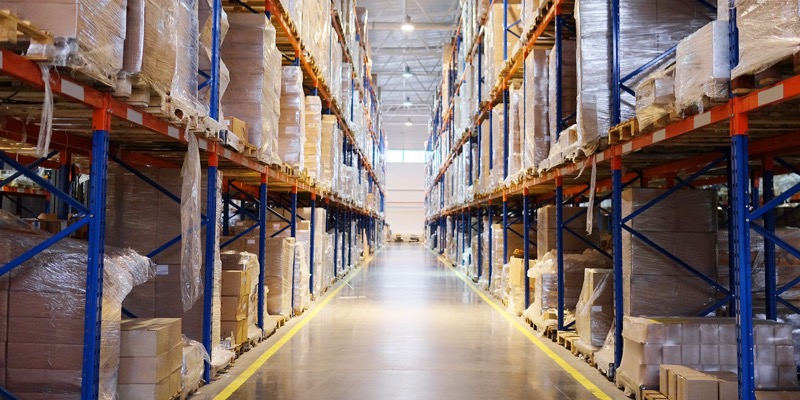 A wide aisle between tall warehouse storage racks filled with palletized goods, boxes, and products, wrapped in plastic for supply chain and inventory management.