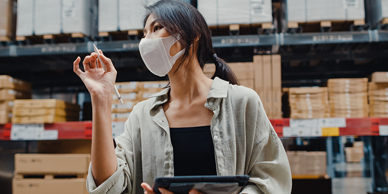 A woman wearing a protective face mask holds a pen and digital tablet while inspecting inventory in a warehouse with shelves stacked with boxes.