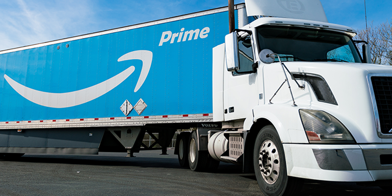 A white Amazon Prime semi truck with a blue trailer featuring the Amazon smile logo and the word 'Prime,' parked outdoors on a clear day.