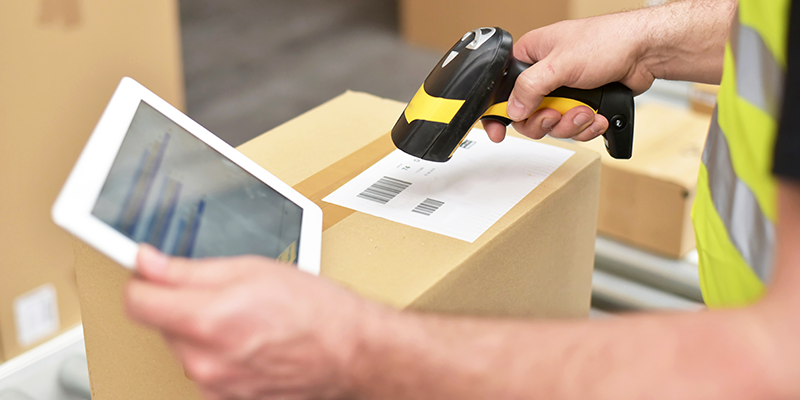 A close-up image of a worker scanning a barcode on a cardboard box using a handheld scanner, while holding a tablet displaying data, representing digital supply chain and inventory management processes.