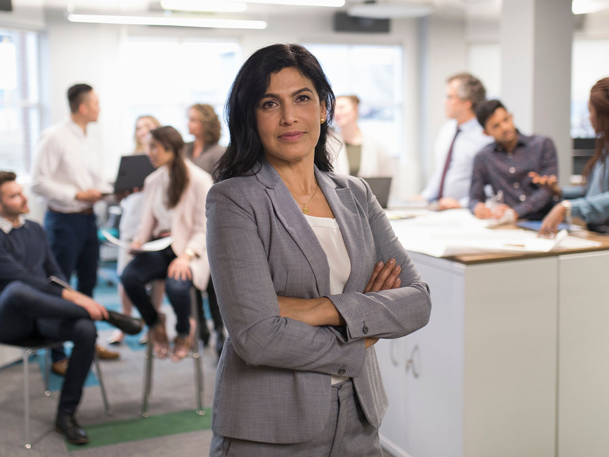 A confident businesswoman in a gray suit stands with arms crossed in the foreground of a modern office, with a diverse group of professionals collaborating in the background.