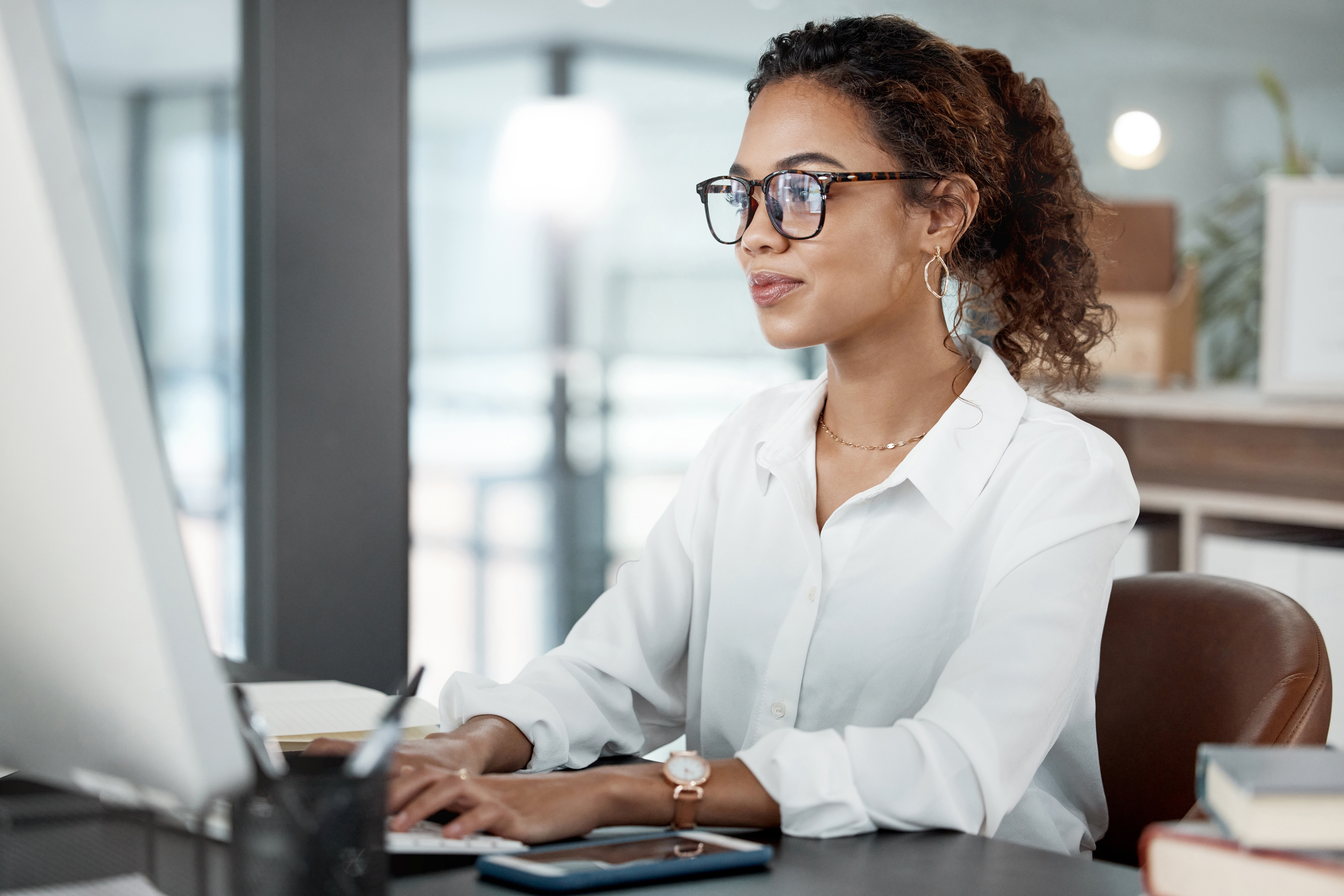 A person wearing a white blouse and gold jewelry, typing on a computer in a modern office setting.