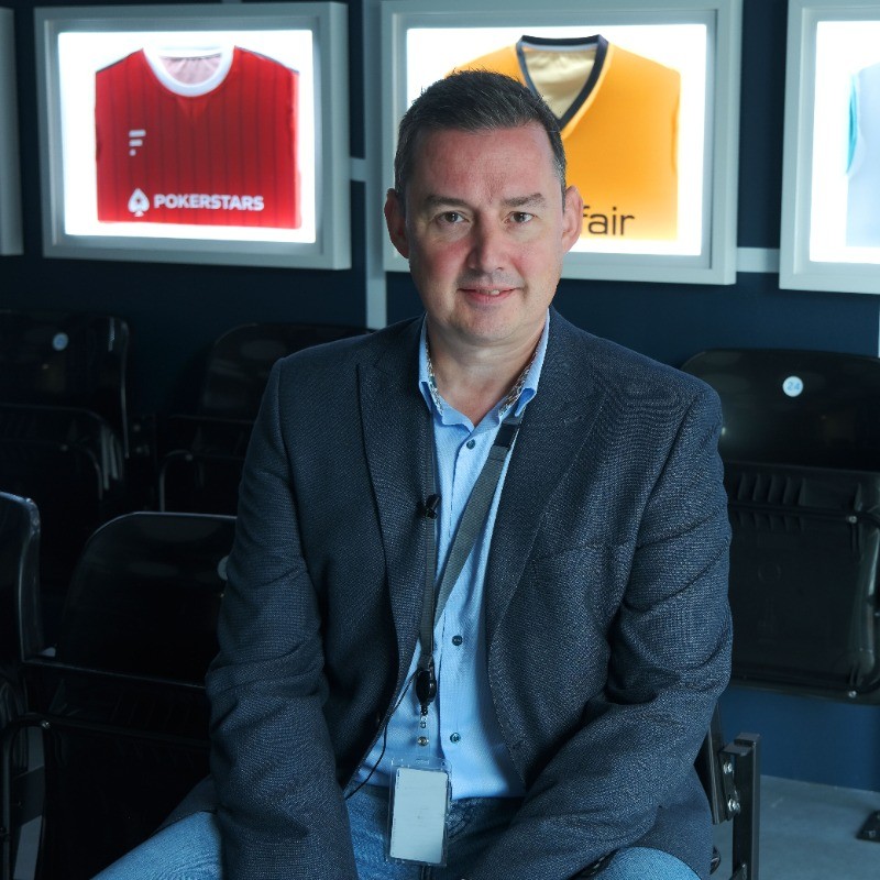 A person seated in front of illuminated framed sports jerseys, wearing a blue shirt and blazer with a lanyard.