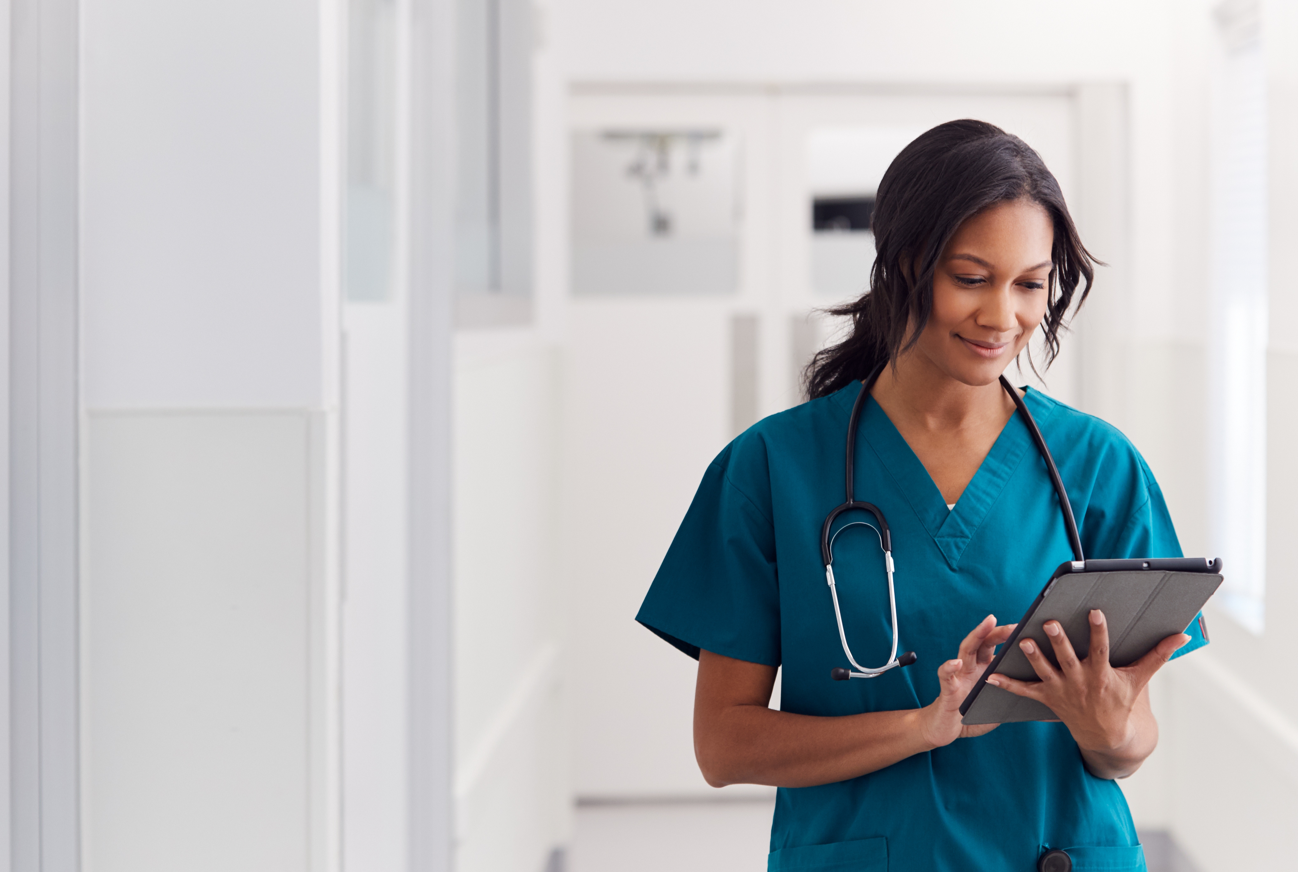 A healthcare professional in scrubs with a stethoscope around her neck uses a digital tablet while standing in a bright hospital hallway.