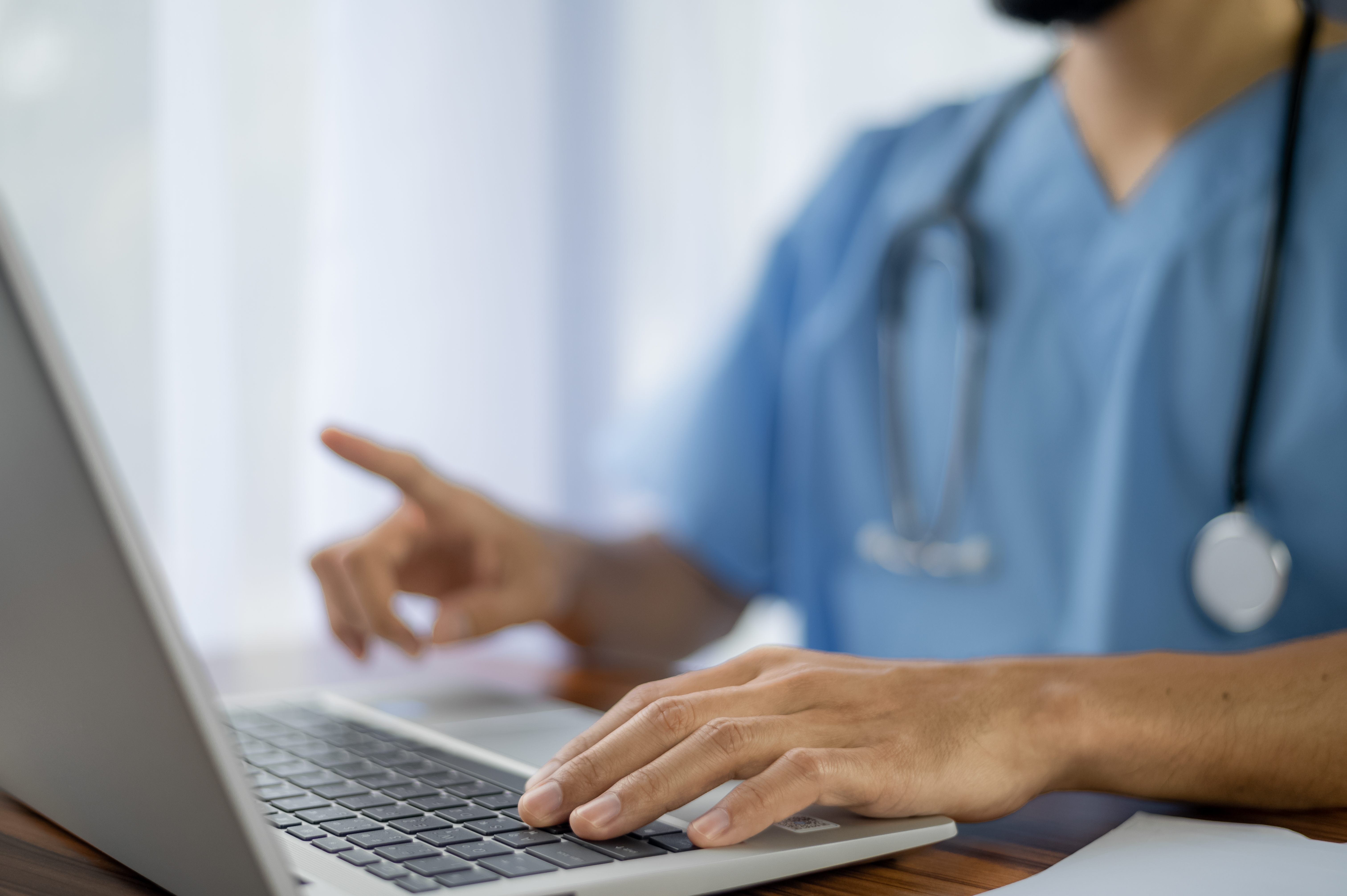 A healthcare professional in blue scrubs with a stethoscope around their neck is working on a laptop, focusing on digital health records or telemedicine tasks.