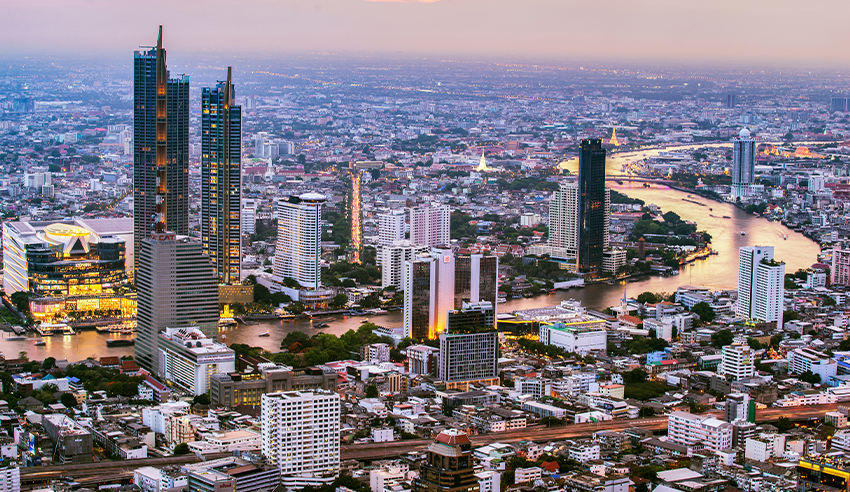 A panoramic view of Bangkok's cityscape featuring modern skyscrapers and buildings along the Chao Phraya River at sunset.