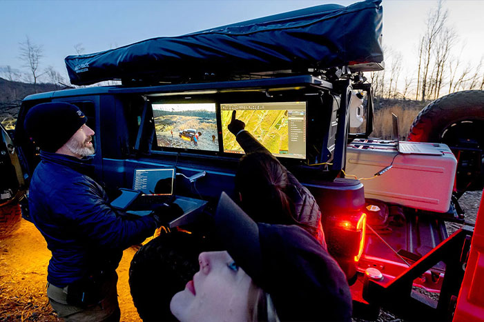 A group of people operating an outdoor mobile command center at night, utilizing advanced technology and computer monitors mounted on a vehicle in a rugged environment.
