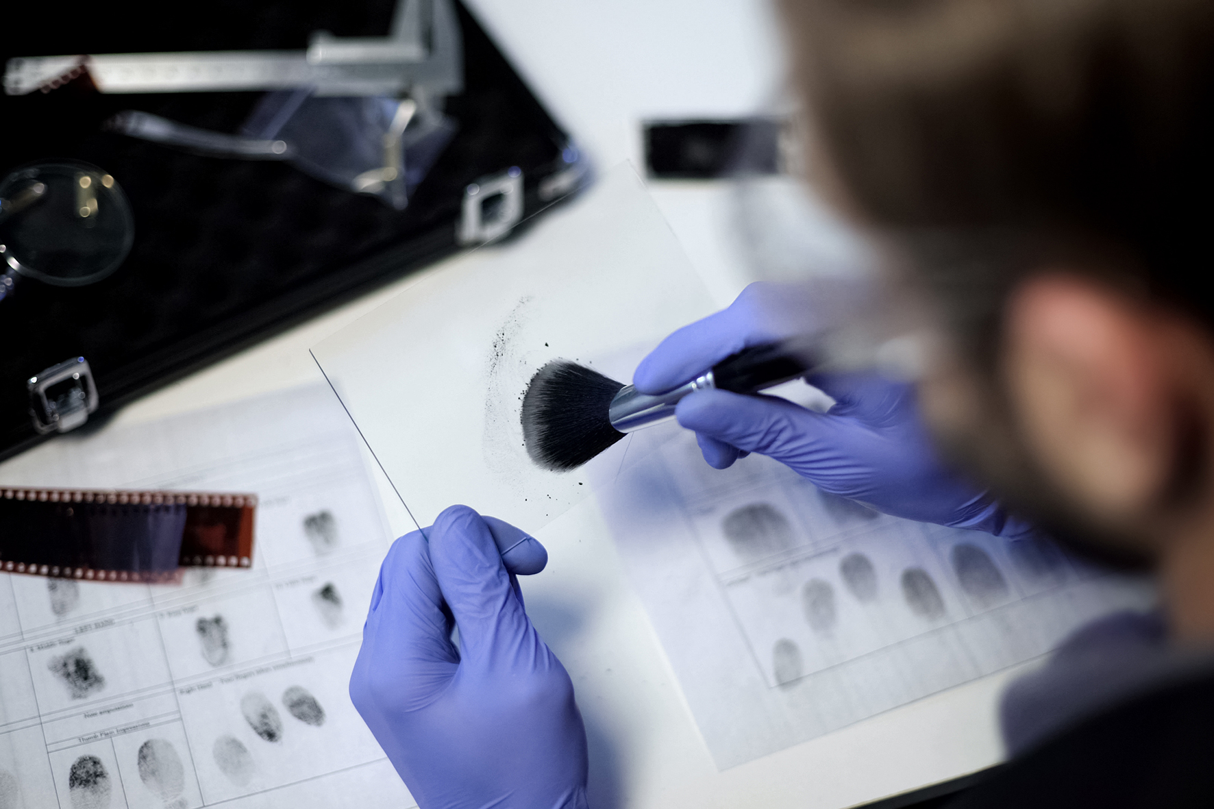 A forensic specialist wearing gloves uses a brush to analyze and collect a fingerprint from evidence, with fingerprint records and analysis tools visible on the desk.