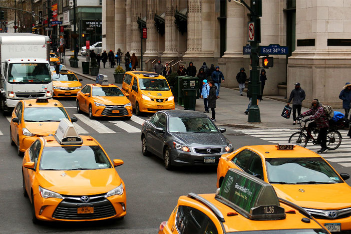 A busy street scene showing multiple yellow taxis and other vehicles in traffic at the intersection of East 34th Street in New York City, with pedestrians and cyclists nearby.