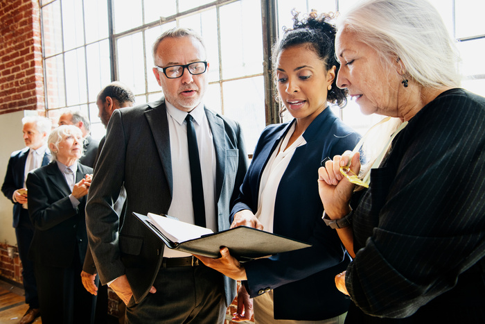 A group of business professionals engaged in discussion, reviewing documents together during a networking event. The scene takes place in a bright, industrial-style venue with large windows.