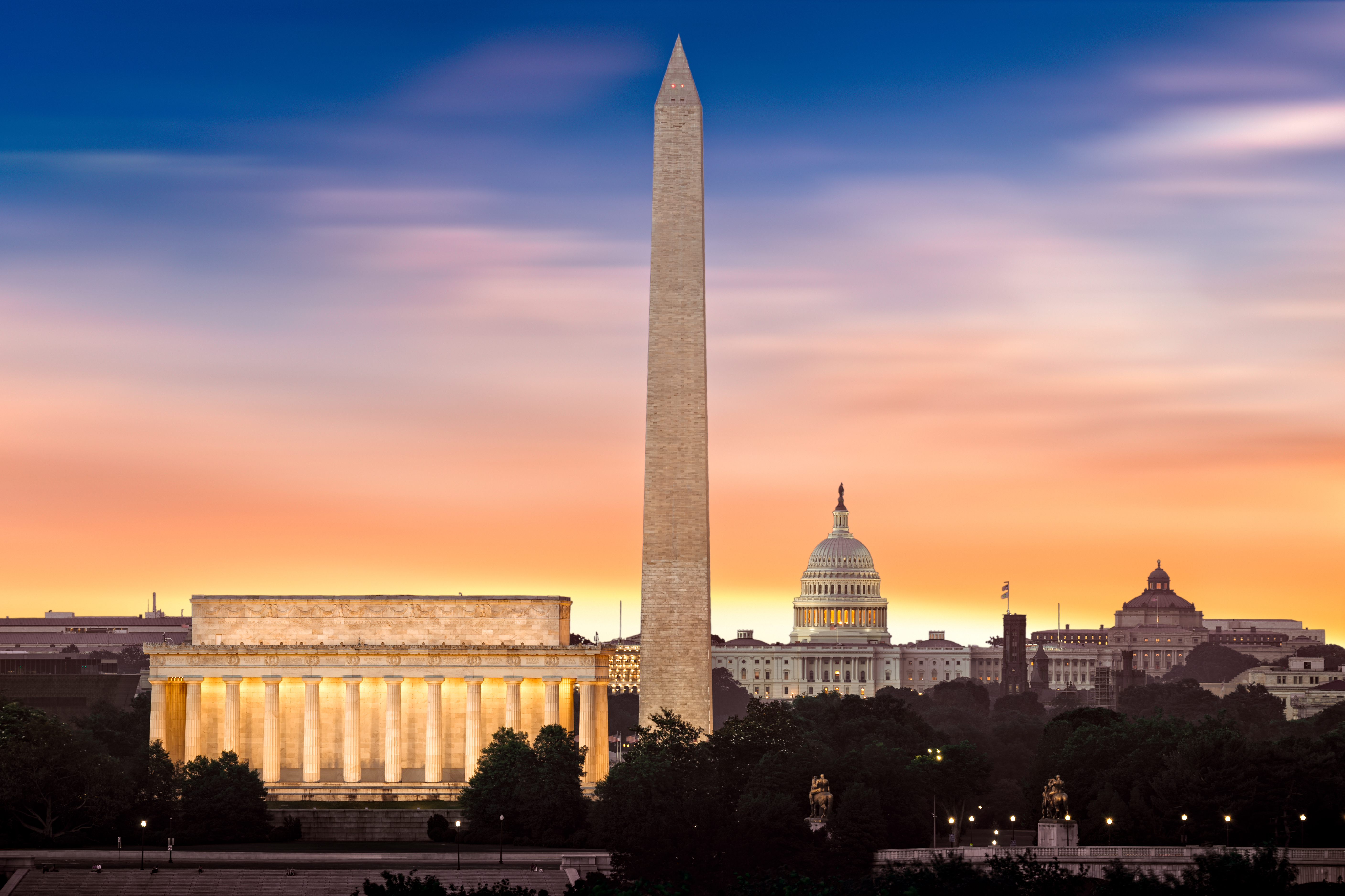 The Lincoln Memorial, Washington Monument, and U.S. Capitol at sunset with a vibrant orange and blue sky.