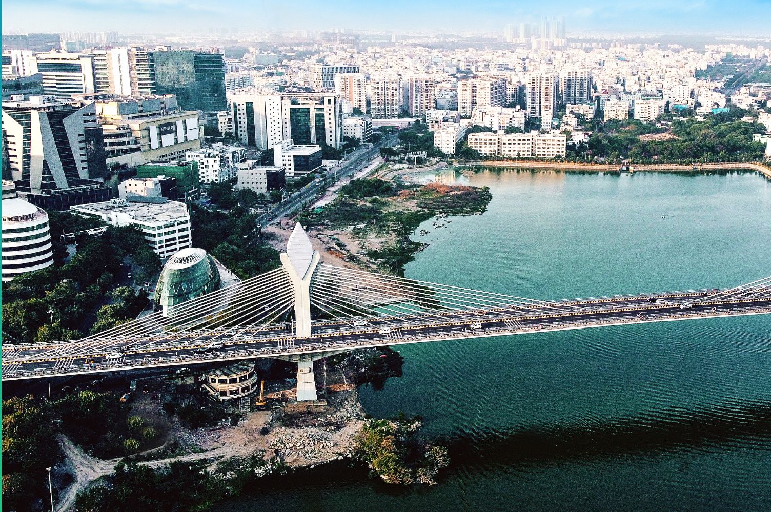 Aerial photograph showcasing a modern cable-stayed bridge spanning over water, with a backdrop of urban buildings, offices, trees, and surrounding cityscape in India.