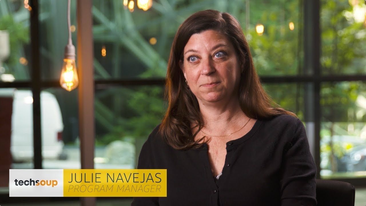 A woman with long brown hair wearing a black top sits in front of a window with greenery outside; text overlay reads "Julie Navejas, Program Manager, TechSoup."
