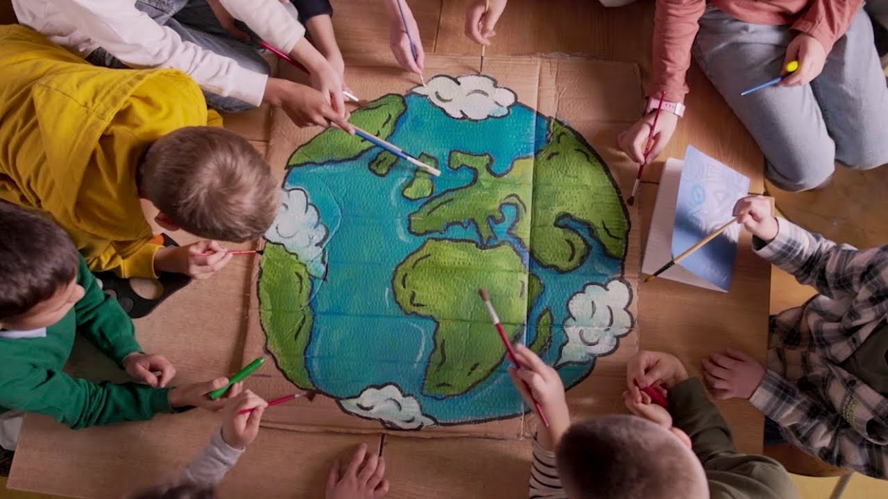 A group of children gathered around a table, collaboratively painting an illustration of the Earth on a large piece of cardboard. The image captures a top-down view, emphasizing creativity, teamwork, and environmental themes.