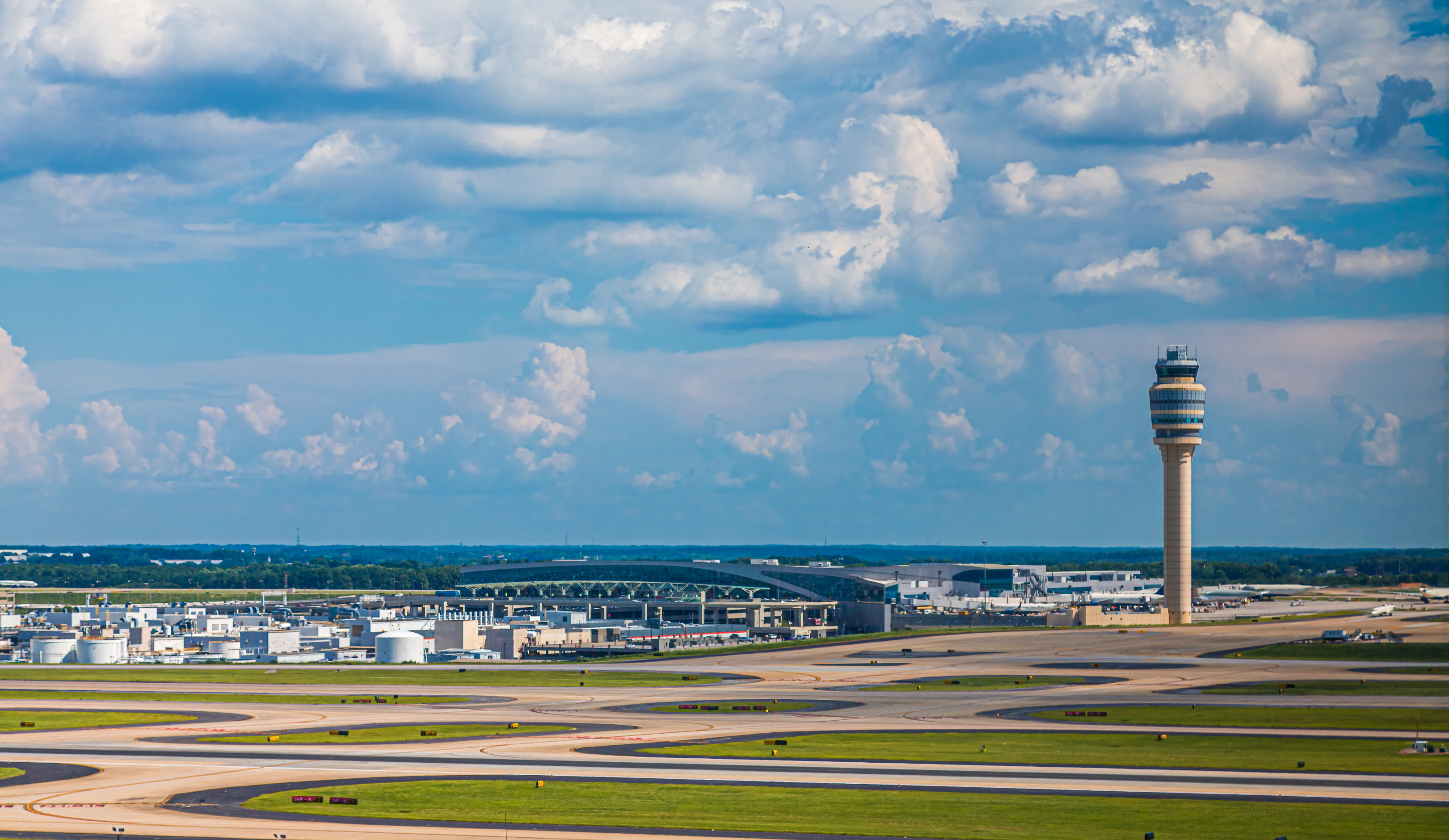 A wide-angle view of an airport showing a prominent air traffic control tower, multiple runways, and airport facilities under a blue sky with scattered clouds.