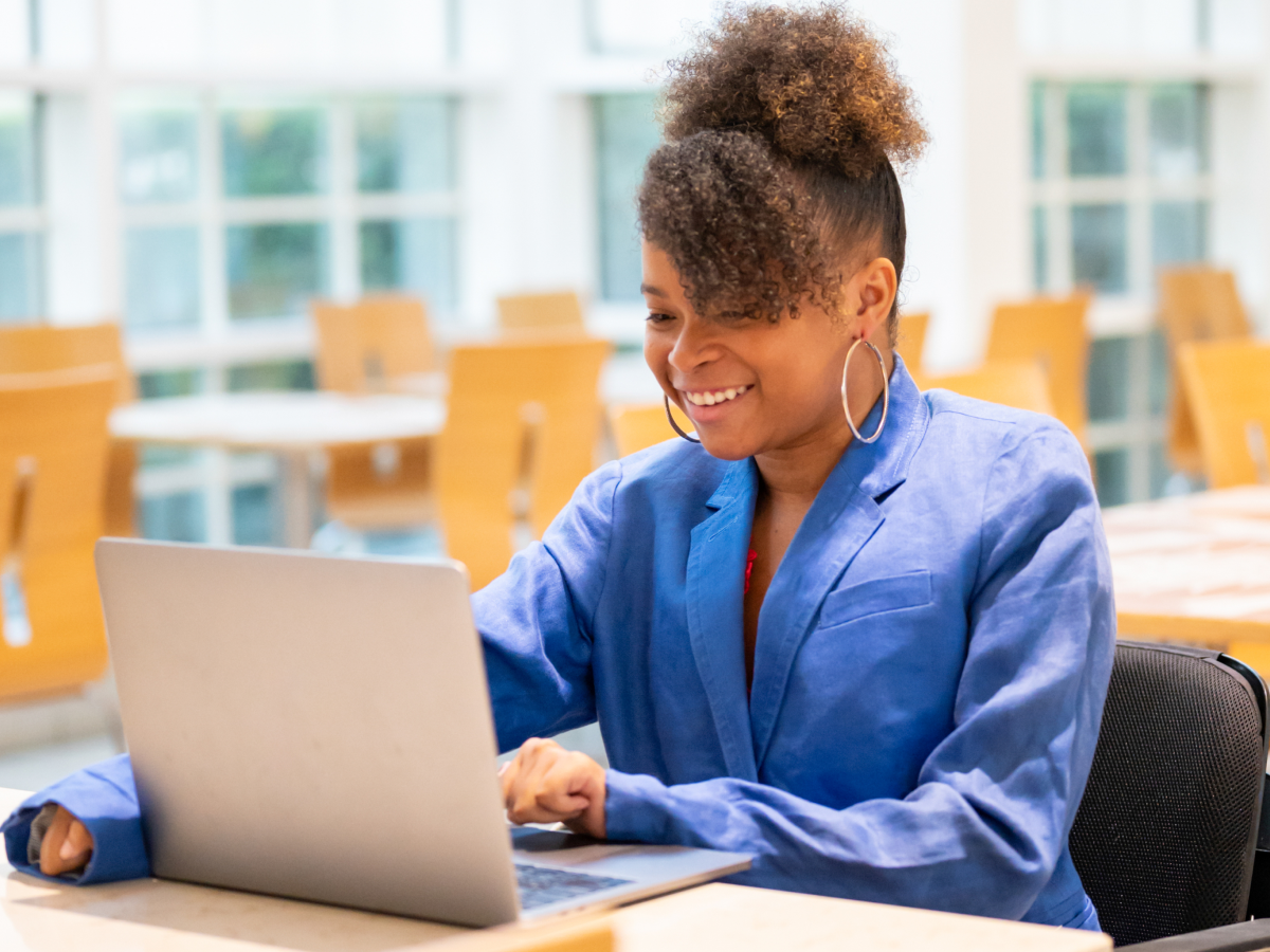 A woman in a blue blazer smiling and working on a laptop in a modern, bright office setting with large windows and wooden chairs in the background.