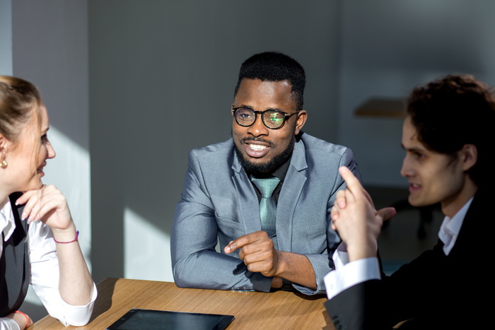 A group of business professionals sitting at a table having a discussion, with one man in a suit and glasses speaking and others listening attentively.