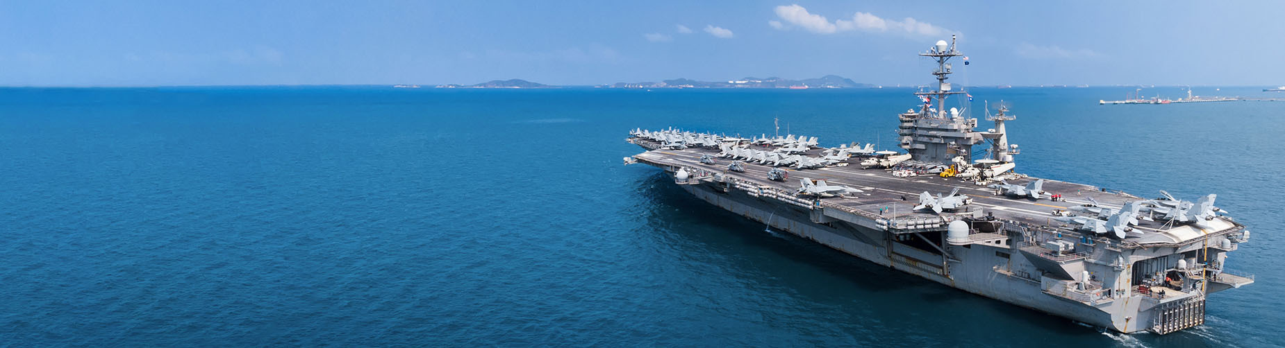 Aircraft carrier with fighter jets docked on a calm blue sea, distant land visible on the horizon.