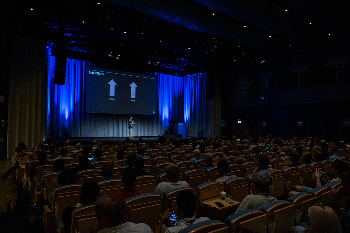 A speaker gives a presentation on stage in a large auditorium filled with an audience. The projection screen shows latency and cost side effects, and the room is illuminated with blue lighting.