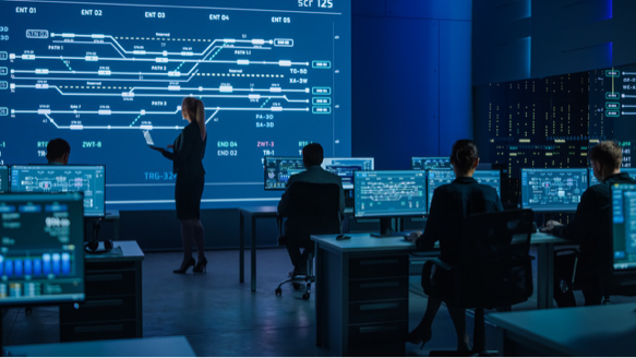 A group of people in a control room with multiple computer screens, while a woman stands pointing at a large digital display showing a complex schematic.