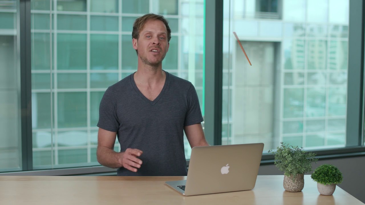 A man stands behind a desk with a laptop and two small potted plants in a modern office setting with large windows and cityscape in the background.
