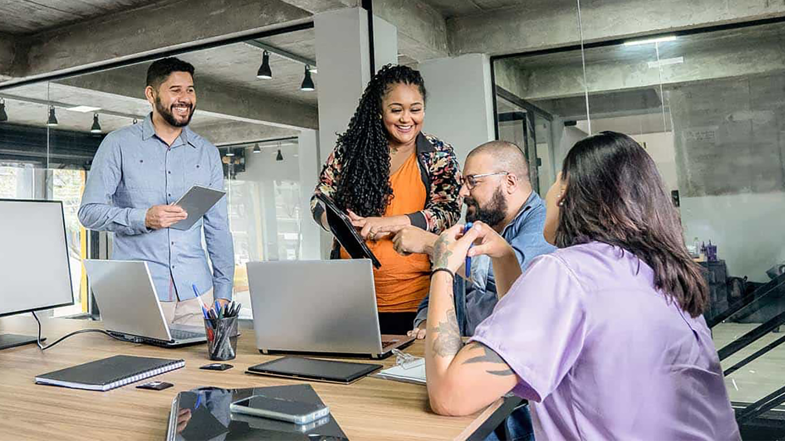 A group of people collaborating around a table with laptops and notebooks in a modern office setting.