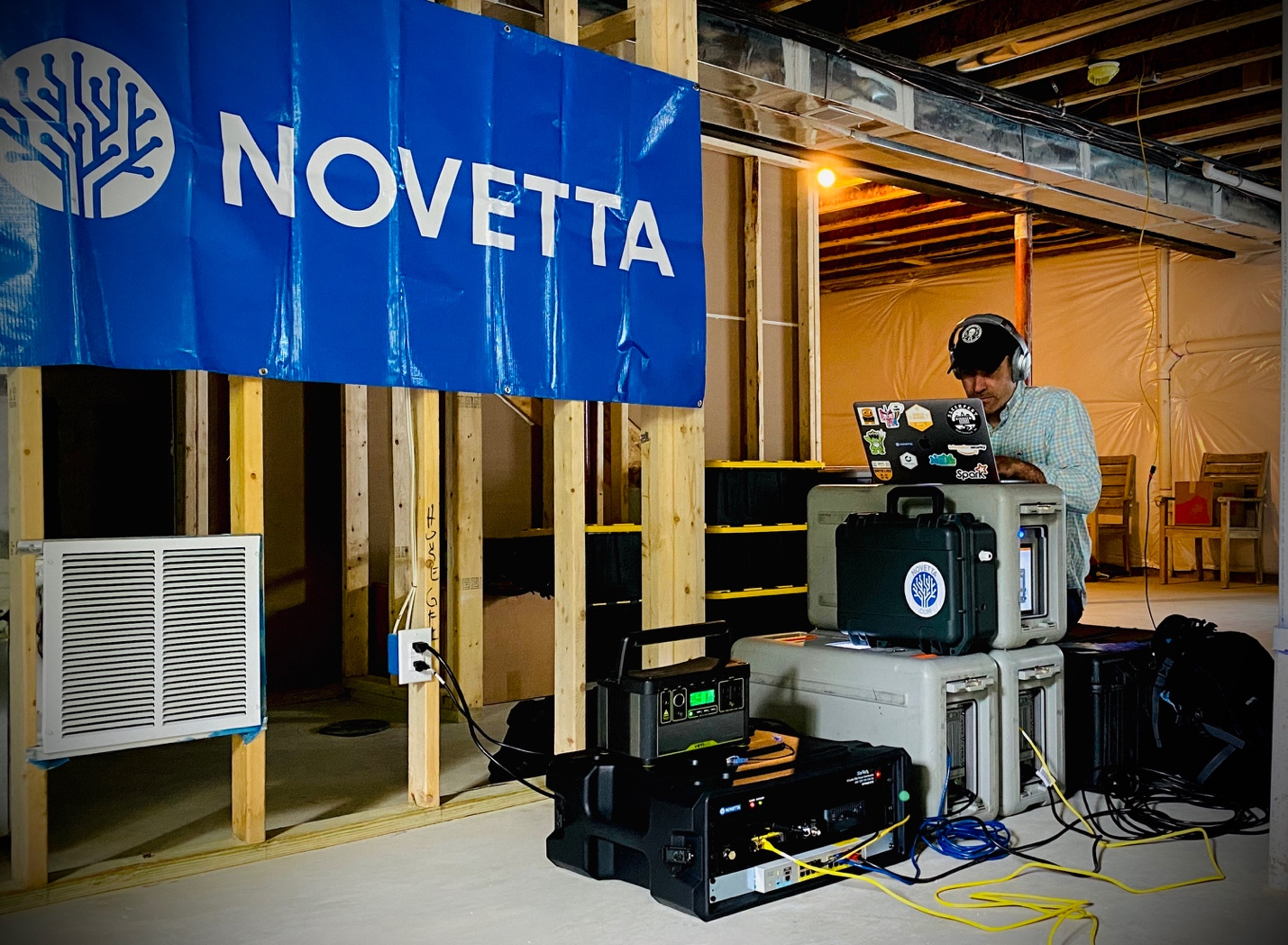 A workspace in a construction-like setting with equipment cases, cables, and a laptop, under a blue "Novetta" banner.