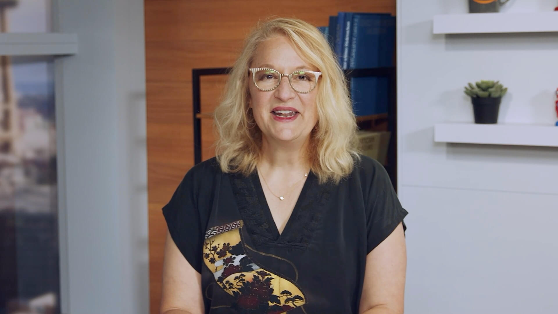 A woman with blonde hair and glasses, wearing a dark blouse, is presenting or speaking in an office setting. The background features shelves with books and a plant, suggesting a business or professional environment.