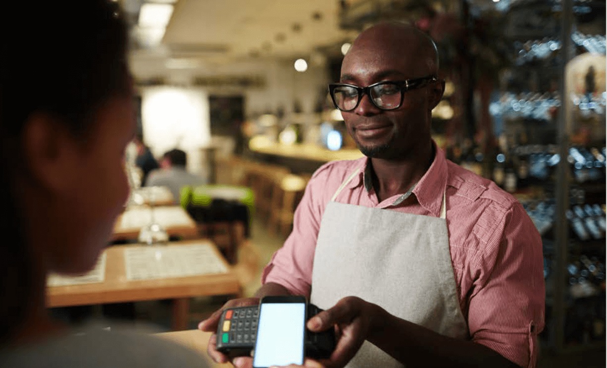 A small business owner wearing an apron processes a payment with a card reader for a customer in a restaurant or cafe setting, illustrating business productivity tools for SMBs.