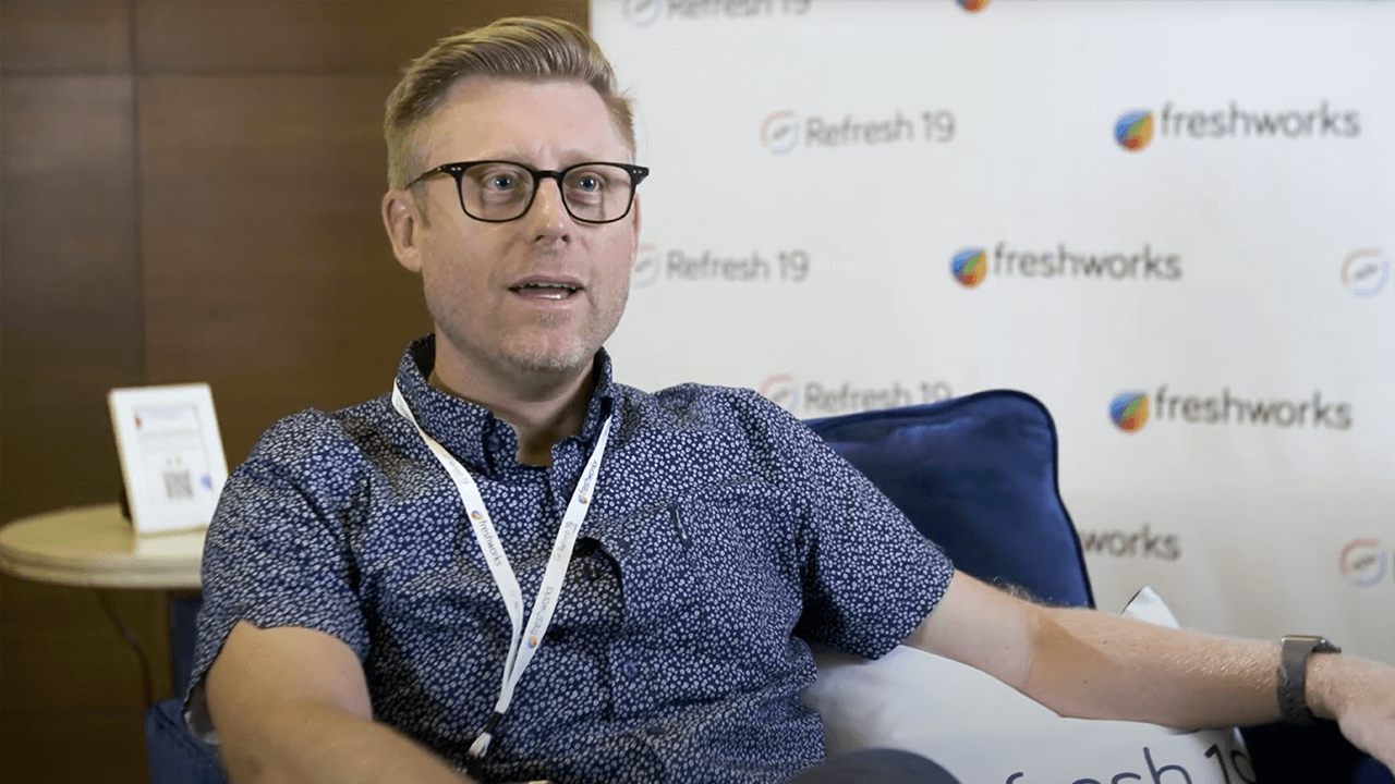 A man wearing glasses sits and speaks during an interview at Refresh 19, with Freshworks branding visible in the background.