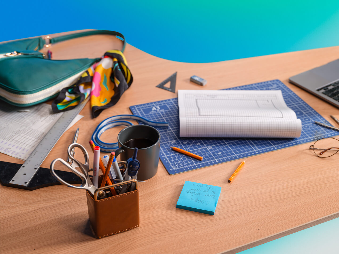 A workspace desk with various supplies including a rolled blueprint, a laptop, ruler, pencils, pens, a cup of coffee, glasses, a purse, and a sticky note, commonly used by architects or designers.