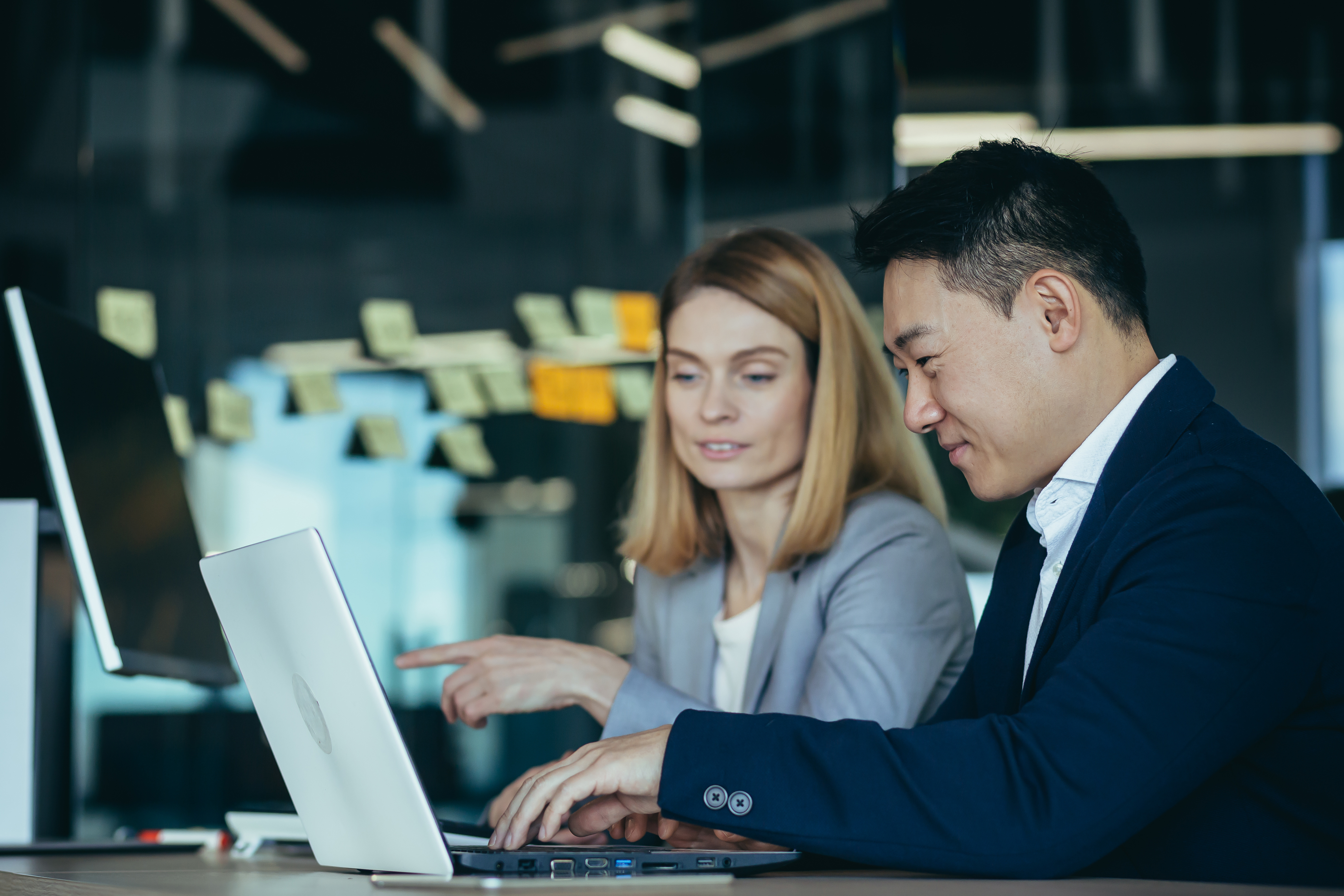 happy coworkers discuss project strategy by looking and pointing at laptop pc computer monitor screen. multiethnic business team in the office. confident mature asian man explaining young female