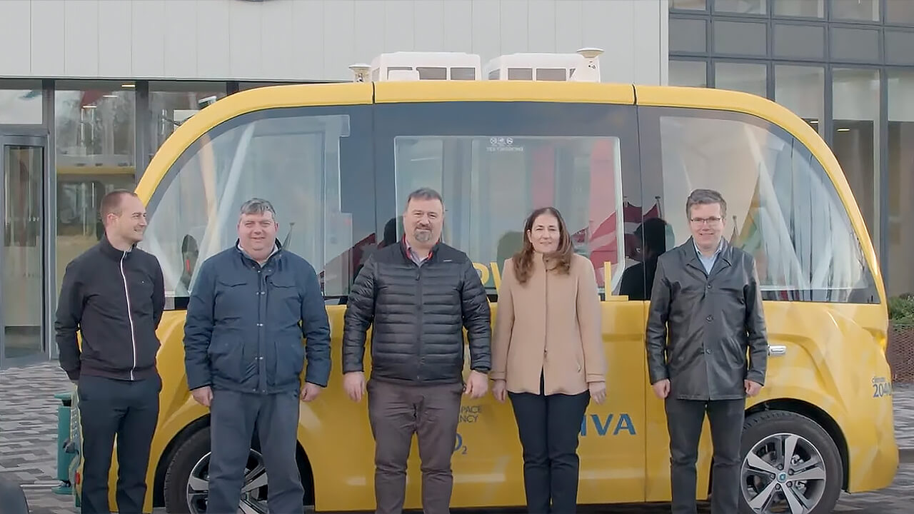 A group of five people standing in front of a modern yellow autonomous shuttle vehicle outside a building.